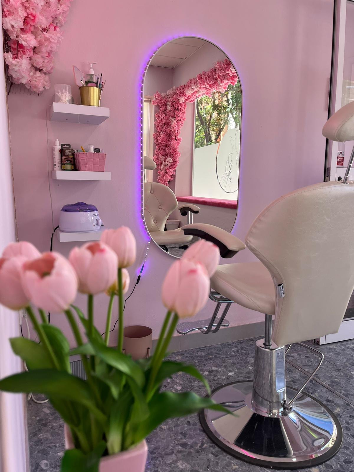 Interior del salón: sillón crema, espejo ovalado con luz morada, flores rosas y estantes con productos
